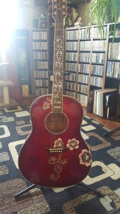 a red guitar sitting on top of a stand in front of a book shelf filled with books