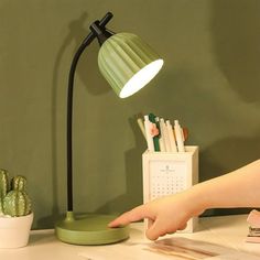 a person pointing at a desk lamp on top of a white table next to a cactus