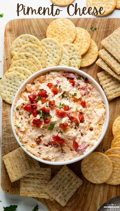 a white bowl filled with dip surrounded by crackers and tomatoes on a cutting board