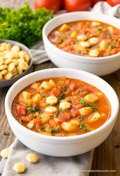 two white bowls filled with soup next to tomatoes and chickpeas on a wooden table