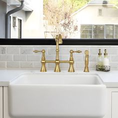 a white kitchen sink with gold faucets in front of a window overlooking the backyard