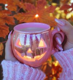 a person holding a pink mug with a glowing pumpkin on it's face in front of fall leaves