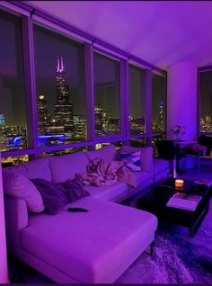 a living room filled with furniture and purple lighting in front of large windows overlooking the city