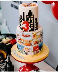 a three tiered cake decorated with comic books and balloons in the shape of city skylines