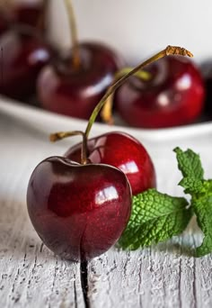 two cherries on a table next to a plate of cherries and mint leaves