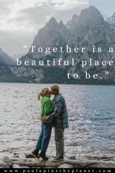 a man and woman standing next to each other on the shore of a lake with mountains in the background