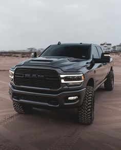 a black ram truck parked on top of a sandy beach next to the ocean and buildings