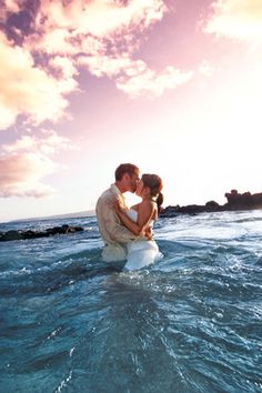 a man and woman kissing in the ocean on their wedding day, while the sun is setting