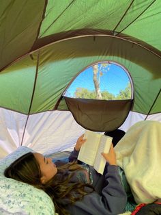 a woman laying in bed reading a book under an open green tent with trees and blue sky reflected in the window