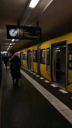 a yellow train pulling into a station with people walking by it and a clock on the wall