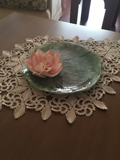 a green plate with a pink flower on it sitting on a lace doily in front of a dining room table