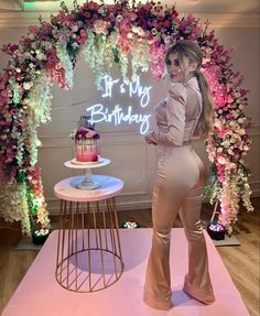 a woman standing in front of a cake on a table with flowers around it and a neon sign that says it's my birthday