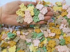 a person holding crocheted flowers in their hand on a white tablecloth with yellow, green and pink colors