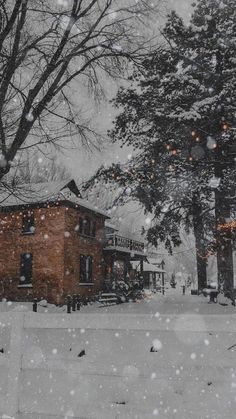 snow falling on the ground and trees in front of a building with lights hanging from it