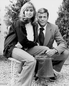 a man and woman sitting on top of a chair in front of some bushes with their arms around each other