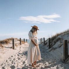 a pregnant woman in a white dress and hat standing on the sand at the beach