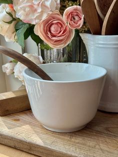 there is a bowl and spoon on the table with flowers in the vase behind it