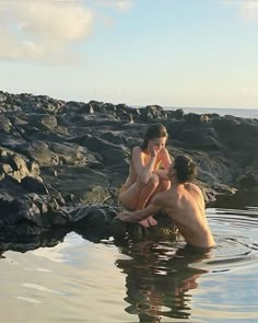 two people are sitting in the water near some rocks and one is holding a cell phone