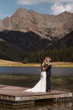 a bride and groom standing on a dock in front of the mountains with their arms around each other