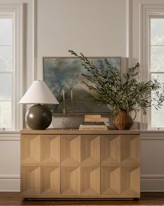 a table with a lamp and vase on it in front of two windows, next to a painting