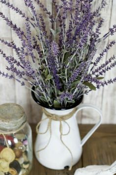 purple flowers are in a white pitcher next to a cookie jar and spoon on a wooden table