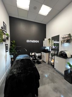 Black accent wall in esthetician room. Lit up sign that says synskn. Green plants accent the room. Marble grey floors and two white walls. Front Yard Flower Bed, Waxing Room, Facial Room