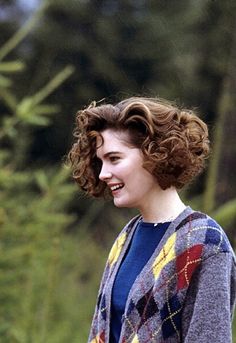 a woman with curly hair standing in front of some trees and smiling at the camera