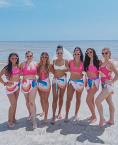 a group of women standing on top of a sandy beach next to the ocean wearing pink and blue bikinis