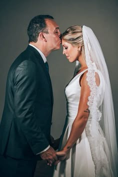 a bride and groom kissing each other in front of a gray background with white lace on the veil