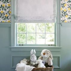 two stuffed animals sitting in a basket next to a window with roman shades on the windowsill