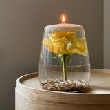 a single yellow rose in a glass vase with gravel around it and a lit candle