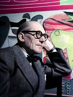 an older man wearing glasses in front of a wall with art on it and a painting behind him