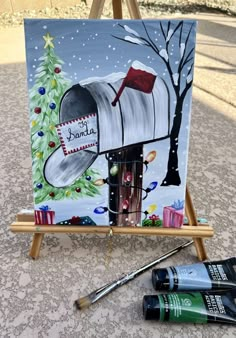 an easel with paint and brushes on it next to a christmas card holder that has a painting of a mailbox in the snow