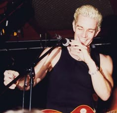 a man with blonde hair holding a guitar and singing into a microphone while standing in front of a microphone