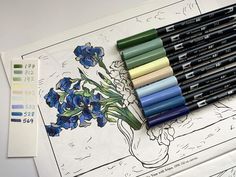 several markers and pencils on top of a drawing sheet with blue flowers in a vase