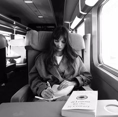 a woman sitting in a train reading a book and writing on a notepad next to a window