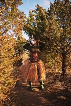 a woman in a costume walking down a dirt road with trees and bushes behind her