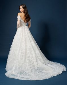 a woman in a white wedding dress with long sleeves and flowers on the skirt is standing against a blue background