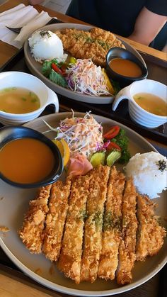 two plates filled with different types of food on top of a wooden table next to bowls of soup and sauces