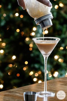 someone pouring something into a martini glass on top of a wooden table in front of a christmas tree