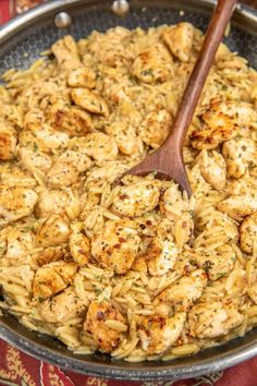 a skillet filled with chicken and pasta on top of a red table cloth next to a wooden spoon