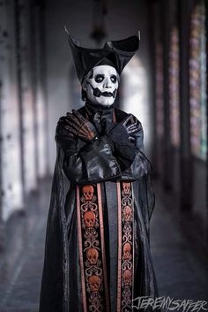 a man in a skeleton mask and long black coat with orange trims is posing for the camera