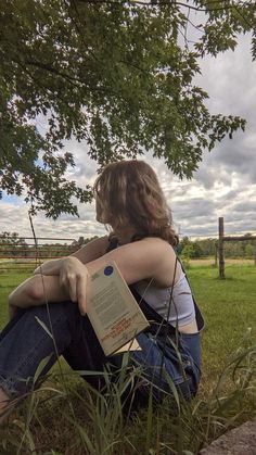 a woman sitting under a tree with her book in her lap and looking at the sky