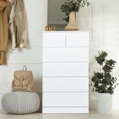 a white chest of drawers sitting next to a plant and a purse on the floor