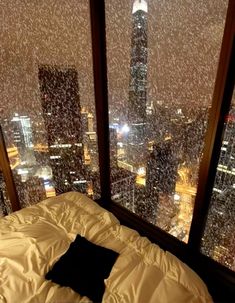 a bed sitting in front of two large windows with city lights on them and snow falling all over the floor