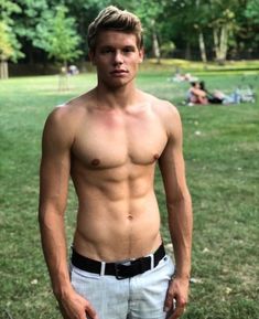 a shirtless man standing in the grass with his hands in his pockets and looking at the camera