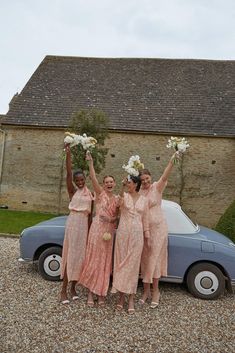 three bridesmaids pose in front of a vintage car with their bouquets raised