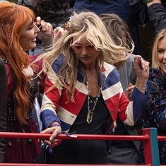 two women standing next to each other in front of a red metal fence with people behind them