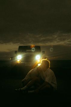 two people sitting on the ground in front of a truck at night with their head lights on