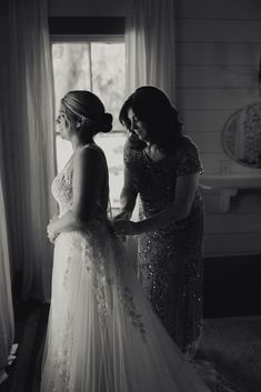 two women standing next to each other in front of a window wearing wedding gowns